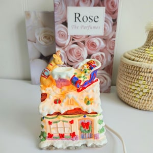 Peut inclure: Figurine de maison de Noël en céramique avec un renne tirant un traîneau rempli de cadeaux. La maison est recouverte de neige et décorée de nœuds rouges et d'une couronne. Un livre avec le mot "Rose" est en arrière-plan.