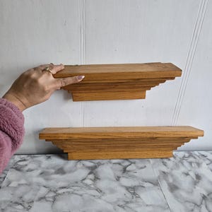 Könnte beinhalten: Zwei Holzregale mit einem dekorativen, geschichteten, gewellten Rand. Die Regale sind hellbraun und an einer weißen Wand montiert.