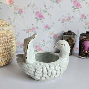 May include: A white ceramic bird-shaped bowl featuring detailed moulded feather textures on the body and wings. The bird has a smooth, curved neck and head with gold-toned accents along the edges of the wings and beak, forming a small open basin.