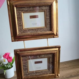 Peut inclure: Deux cadres photo dorés suspendus à une corde, chacun avec un cadre intérieur plus petit. Les cadres sont reliés par une corde et ont des glands décoratifs. Les cadres sont étiquetés "Treasured Memories" et mesurent 9 x 13 cm.