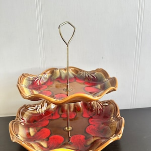 May include: Two-tiered ceramic serving stand with a brown and red swirl pattern. The stand has a scalloped edge and a metal rod connecting the two tiers.