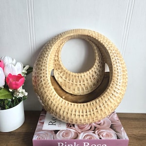 May include: A woven, light brown decorative basket with a circular design. The basket sits atop a book titled "Pink Rose" with a pink cover. A small, pink hand sculpture in the shape of a heart is in front of the book.
