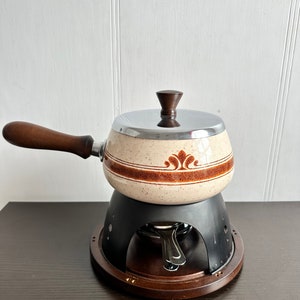 Puede incluir: Una olla de fondue vintage con una olla de cerámica marrón y blanca con un diseño floral marrón, un soporte de metal negro y una base de madera.