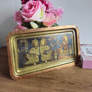 Peut inclure: Un plateau rectangulaire en laiton avec un bord en cuivre présente un motif gravé de trois figures de style égyptien antique. Les figures sont représentées en or et en noir sur un fond bleu foncé.
