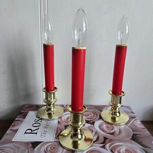 Könnte beinhalten: Drei elektrische Kerzen mit roten Schäften und klaren Glühbirnen, in goldfarbenen Haltern. Die Kerzen stehen auf einem Buch mit dem Text "Pink Rose" und einem floralen Muster.