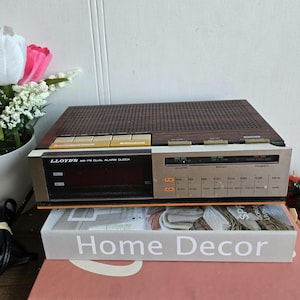 Puede incluir: Un reloj despertador dual AM/FM LLOYD'S vintage con una parte superior de veta de madera marrón y un panel frontal plateado. El reloj tiene una pantalla LED e indicadores de frecuencia de radio. El texto "Home Decor" es visible en un libro.