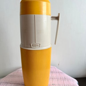 Può includere: Un thermos giallo e bianco con un design a coste e un manico. Il thermos ha la scritta "THERMOS" stampata sul lato.