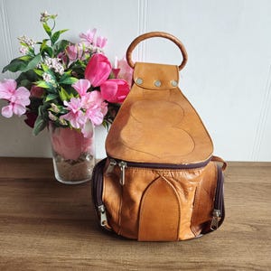 May include: A brown leather backpack with a unique design. The bag has a top handle and two zipped compartments.