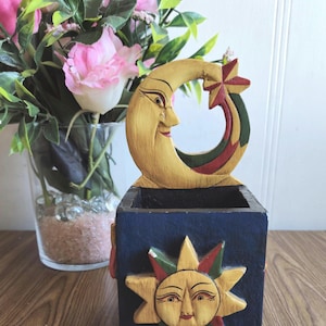 Könnte beinhalten: Eine Holzkiste mit einem geschnitzten Halbmond und Stern oben und einer geschnitzten Sonne auf der Vorderseite. Die Kiste ist in Blautönen, Gelb, Rot und Grün bemalt.