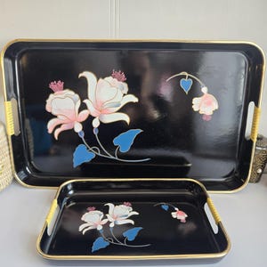 May include: Two black serving trays with gold trim and woven handles. The trays feature a floral design with pink and white flowers and blue leaves. The larger tray is rectangular, and the smaller tray is also rectangular.