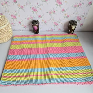 Peut inclure: Un tapis rectangulaire multicolore à rayures horizontales roses, oranges, jaunes et bleues. Le tapis a un bord frangé et est posé sur une surface blanche. Des bougeoirs décoratifs et un panier en osier sont en arrière-plan.