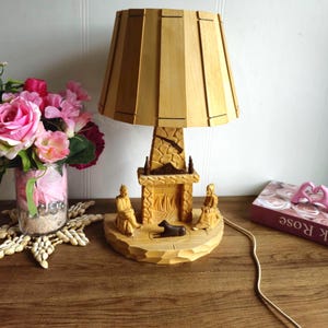 Könnte beinhalten: Eine Holzlampe mit einer geschnitzten Kamin-Szene. Die Lampe hat einen braunen Holzschirm und einen Sockel, der einen Kamin mit zwei Figuren und einem Hund darstellt. Die Lampe steht auf einem Holztisch mit einem rosa Buch und einer Vase mit Blumen.