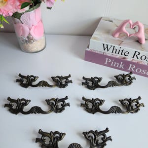 May include: Six antique bronze drawer pulls with ornate, scrolling designs. The pulls are arranged on a white surface, showcasing their intricate details and aged finish. A vase of pink flowers and decorative books are in the background.