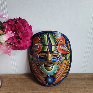 Op de afbeelding: Een kleurrijk handgeschilderd houten masker met een menselijk gezicht, vogels en bloemmotieven. Het masker is ovaalvormig en heeft een donkerblauwe achtergrond.