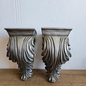 Op de afbeelding: Twee zilverkleurige wandkandelaars met een klassiek ontwerp. Elke kandelaar heeft een rechthoekige bovenkant en een decoratief, vloeiend, bladvormig patroon. De kandelaars worden getoond tegen een witte muur en een houten oppervlak.