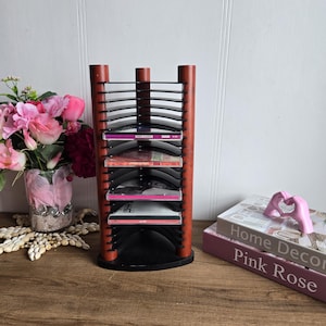 Peut inclure: Une tour à CD en bois avec une base noire et 10 fentes pour CD. La tour est remplie de CD, dont certains sont visibles. La tour est posée sur une table en bois.