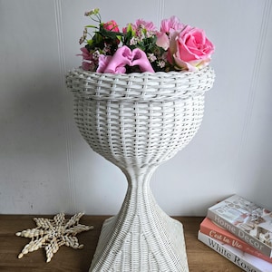 Puede incluir: Un soporte de planta de mimbre blanco con una base cuadrada y una parte superior en forma de jarrón. El soporte está lleno de flores rosas.