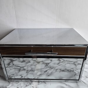 Op de afbeelding: Een broodtrommel van chroom met een lade van houtlook en een spiegelende voorkant. De broodtrommel staat op een wit marmeren oppervlak.