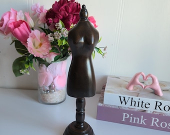 May include: A dark brown wooden dress form with a round base and a decorative top. The dress form is displayed on a white surface, with a vase of pink and burgundy flowers in the background.
