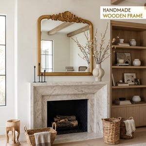 Könnte beinhalten: Ein goldgerahmter Spiegel mit Verzierungen hängt über einem Marmorkamin. Der Raum verfügt über ein Bücherregal aus Holz, eine weiße Vase mit Zweigen und Weidenkörbe. Der Text "HANDMADE WOODEN FRAME" ist sichtbar.