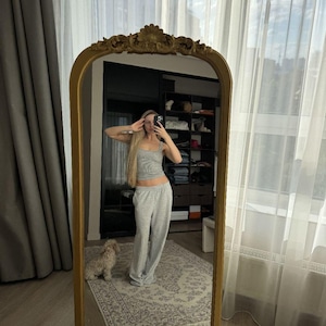 Può includere: Un alto specchio da terra ad arco con una cornice dorata ornata da intagli decorativi sulla parte superiore e inferiore. La superficie in vetro riflettente dello specchio mostra un top senza maniche grigio chiaro e pantaloni da casa coordinati.