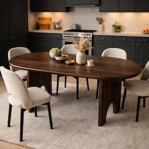 Puede incluir: Una mesa de comedor ovalada de color marrón oscuro con cuatro sillas. La mesa es de madera y tiene una superficie lisa. Las sillas tienen asientos de tela de color claro y patas negras. La mesa está puesta con un jarrón, fruta y una taza de café.