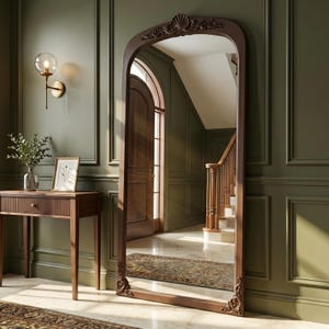 Puede incluir: Un espejo grande y arqueado con un marco de madera marrón oscuro y tallas decorativas. El espejo refleja un pasillo con una escalera y una alfombra estampada. El espejo está apoyado contra una pared verde.