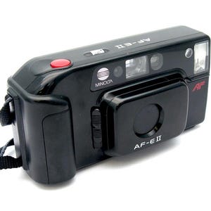 Op de afbeelding: Een zwarte Minolta AF-E II point-and-shoot camera met een rode knop bovenop en een zwarte band.