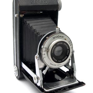 May include: A vintage Kodak folding camera with a black leather-textured body and a black bellows. The silver lens assembly features the text Kodak Anastar 100 mm f/6.3, London, and Dakon Shutter. The camera has a metal frame and a top handle.
