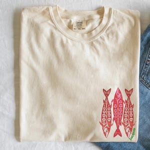 Distressed oversized graphic tee with a vintage fisherman aesthetic. Small red sardines graphic on the front chest, full-size distressed red sardines print on the back with sardines in green script. Nautical, retro inspired design with a worn-in look