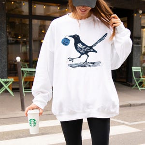 May include: White sweatshirt featuring a black and navy blue graphic of a bird carrying a blue pumpkin. The bird is walking on a navy blue line. The sweatshirt has long sleeves.
