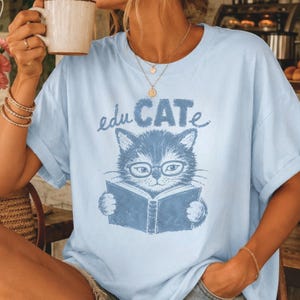 May include: A light blue t-shirt featuring a dark blue graphic print of a cat wearing glasses and reading an open book. The text above the cat reads "eduCATe" in a stylized font. The shirt has a crew neckline and short sleeves.
