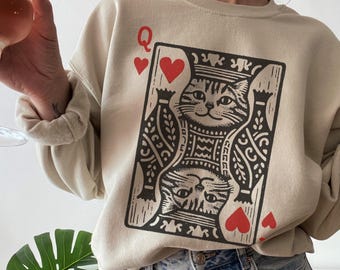 May include: Beige sweatshirt featuring a playing card design with a cat illustration. The card shows the Queen of Hearts, with red hearts and the letter "Q". The sweatshirt is paired with blue jeans.