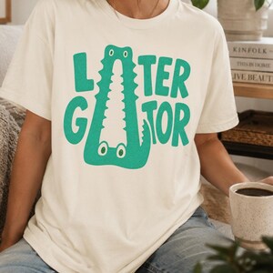 IVORY Comfort Colors graphic tee featuring a playful Later Gator alligator illustration forming the letter A in chunky retro green typography.