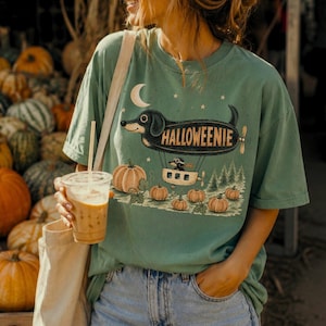 May include: A sage green short-sleeve t-shirt featuring a graphic of a dachshund shaped like a blimp with the text HALLOWEENIE. The design includes a crescent moon, stars, a small basket with a dog, and several orange pumpkins on the ground.