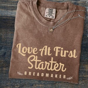May include: A brown t-shirt with the text "Love At First Starter" and the words "Breadmaker" below it. The shirt has a distressed look and is made of a soft, comfortable material.