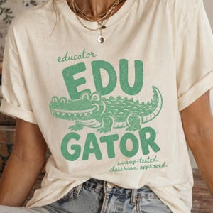 May include: A cream-coloured t-shirt featuring a green graphic of an alligator. The text reads educator, EDU GATOR, and swamp-tested, classroom approved. The design has a distressed texture. The shirt has short sleeves and a crew neckline.