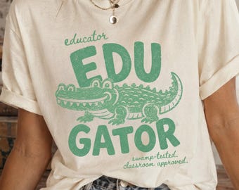May include: A cream-colored t-shirt featuring a green graphic of an alligator. The text reads educator, EDU GATOR, and swamp-tested, classroom approved. The design has a distressed texture. The shirt has short sleeves and a crew neckline.