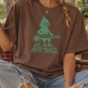 May include: A dark brown short-sleeve t-shirt featuring a green graphic of a bird sitting on a tree stump playing a stringed instrument. Below the bird, the text reads STAY TUNED in a stylized green font. The shirt has a crew neckline.