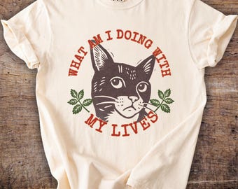 May include: Cream-colored t-shirt featuring a distressed graphic of a cat's face with the text "WHAT AM I DOING WITH MY LIVES" in a circular design. The cat is dark brown with green leaves on either side. The shirt has short sleeves.