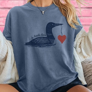 May include: A blue t-shirt with a loon design holding a red heart. The text "a little loon" is printed above the loon. The shirt has a relaxed fit and short sleeves.