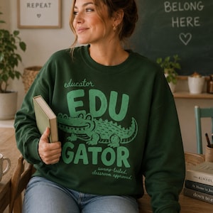 May include: A dark green long-sleeve sweatshirt featuring a light green graphic of an alligator and the text "educator EDU GATOR swamp-tested classroom approved." The sweatshirt has a crew neckline and ribbed cuffs, worn with blue denim jeans.