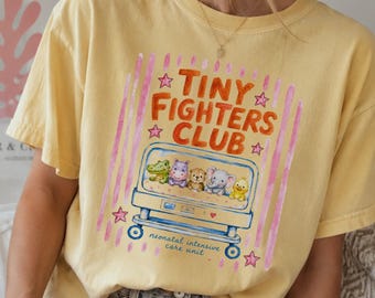 May include: A pale yellow t-shirt featuring a graphic print with the text Tiny Fighters Club and Neonatal Intensive Care Unit. The design includes a blue hospital bassinet containing a cartoon alligator, hippo, bear, elephant, and chick.
