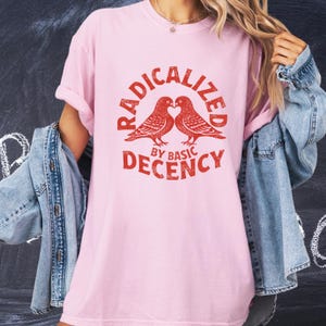 May include: A light pink t-shirt with the words "RADICALIZED BY BASIC DECENCY" in red, along with a graphic of two red doves facing each other. The shirt has short sleeves and a crew neck.
