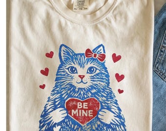 May include: Cream-colored t-shirt featuring a blue cat with a red bow and a heart-shaped graphic that says "BE MINE." The design includes red hearts scattered around the cat. The shirt has a crew neck and short sleeves.