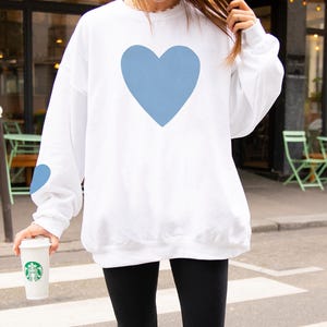 White crewneck sweatshirt featuring a large Carolina blue heart on the chest and a smaller matching Carolina blue heart on the right sleeve.