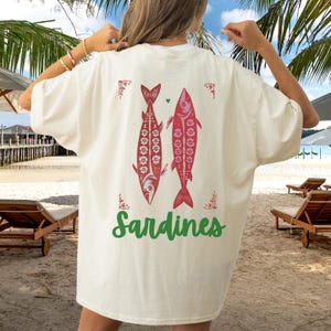 Distressed oversized graphic tee with a vintage fisherman aesthetic. Small red sardines graphic on the front chest, full-size distressed red sardines print on the back with sardines in green script. Nautical, retro inspired design with a worn-in look