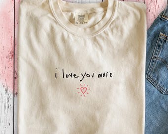 May include: A beige t-shirt with the text "i love you more" written in black ink and a small red heart with rays of light around it.