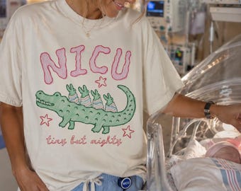 May include: A cream-colored short-sleeve t-shirt featuring a graphic of a green alligator carrying three small figures on its back. The text NICU is printed in pink above the alligator, and the phrase tiny but mighty is printed in pink below.