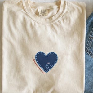 May include: Cream-colored t-shirt with a blue heart-shaped patch. The patch has a white stitched outline and the number "250" inside. The word "Semiquincentennial" is written in red cursive along the heart's edge. The shirt is folded.